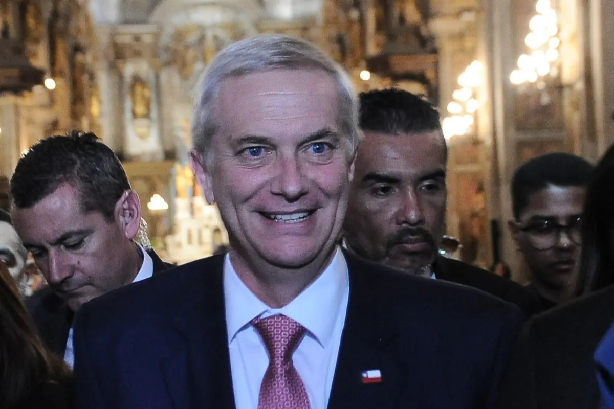 Hombre de cabello canoso con traje oscuro y corbata roja sonriendo entre otras personas en un interior iluminado y elegante