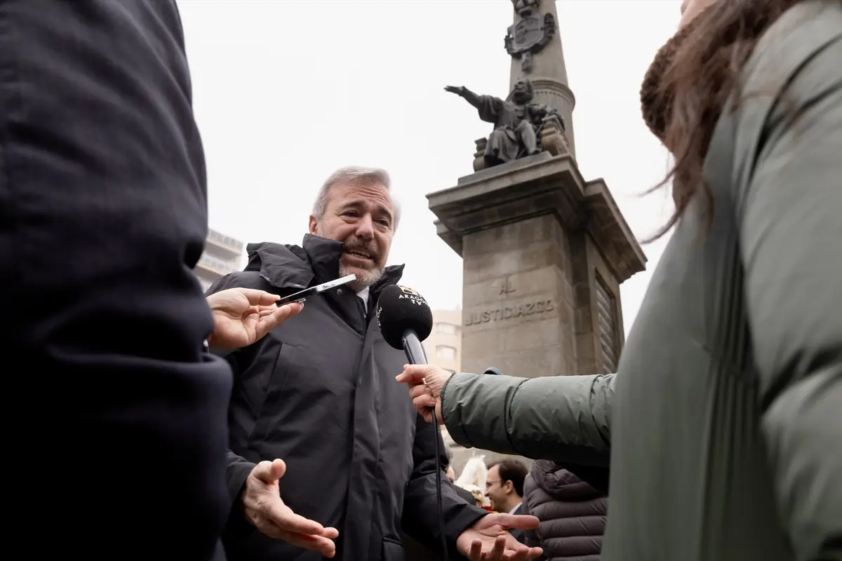 Hombre con abrigo oscuro atiende a varios micrófonos de periodistas frente a un monumento de piedra en una plaza urbana