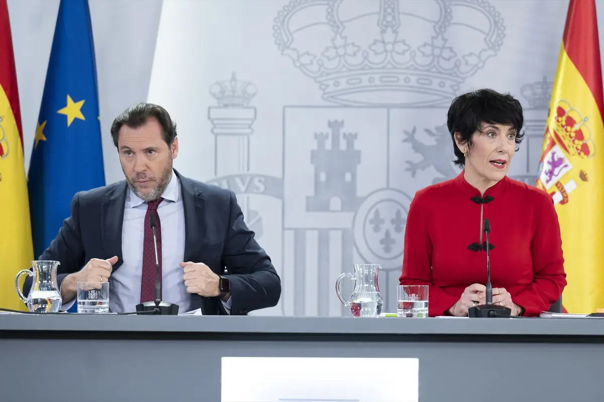 Dos portavoces del gobierno español sentados en una rueda de prensa oficial con banderas de España y de la Unión Europea al fondo