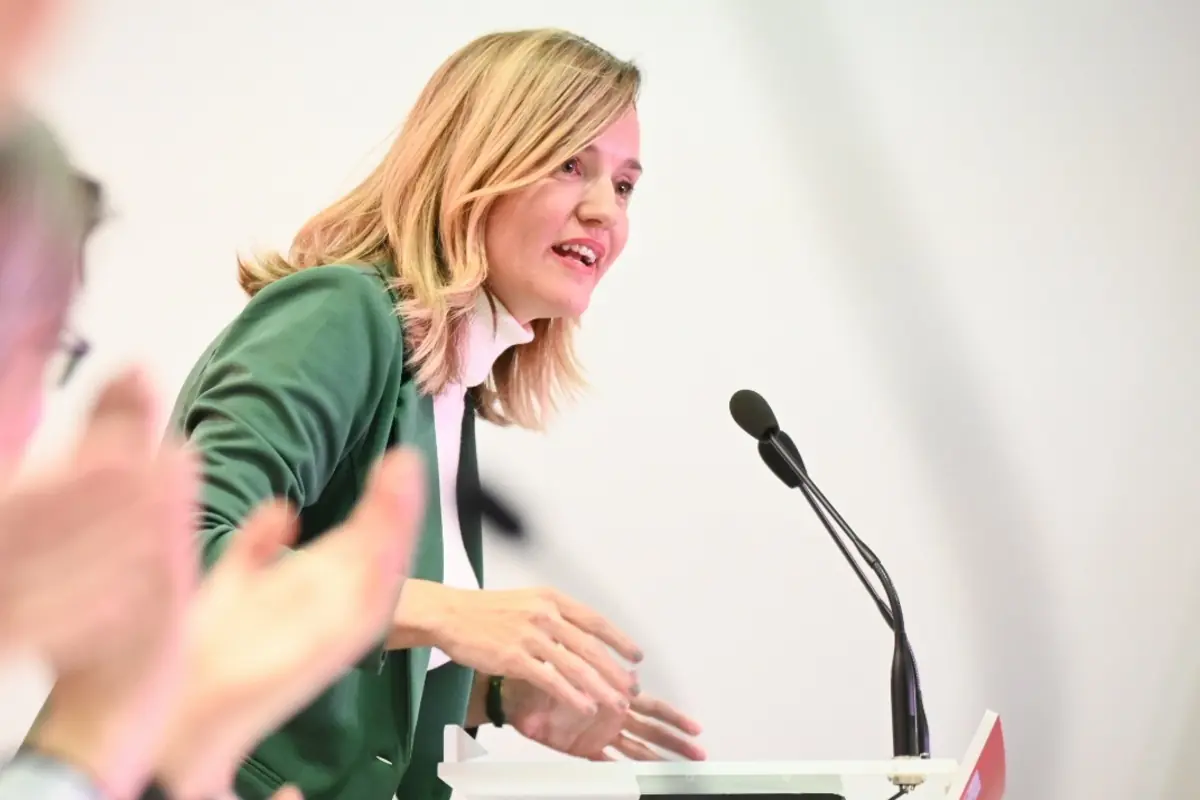 Mujer rubia con chaqueta verde hablando en un atril con micrófono mientras el público aplaude