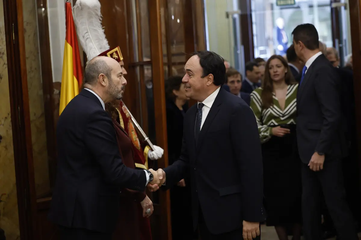 Dos hombres vestidos de traje se dan la mano en un entorno formal con una bandera de España y una persona vestida con uniforme ceremonial al fondo, mientras varias personas conversan y caminan en el lugar.