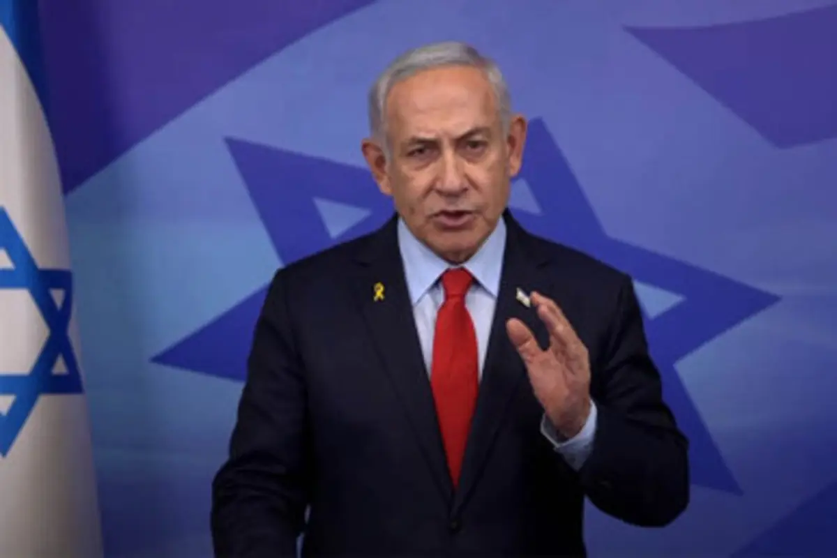 Hombre de cabello canoso con traje oscuro y corbata roja hablando frente a un fondo azul con la bandera de Israel