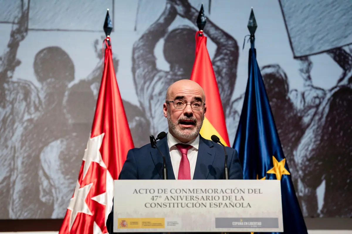 Hombre con gafas y barba hablando en un podio durante un acto de conmemoración del 47 aniversario de la Constitución Española con banderas de fondo