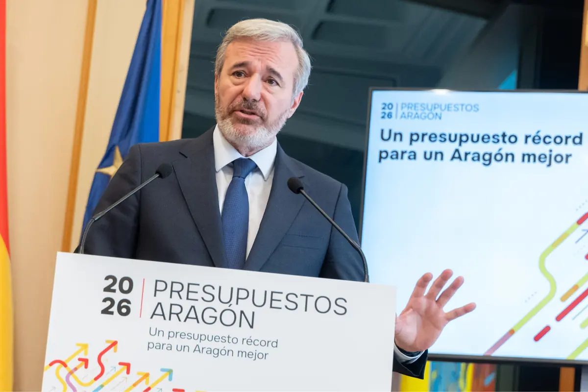 Hombre de cabello canoso y barba hablando en una conferencia de prensa sobre los presupuestos de Aragón 2026 con un cartel y una pantalla de fondo que muestran el lema Un presupuesto récord para un Aragón mejor