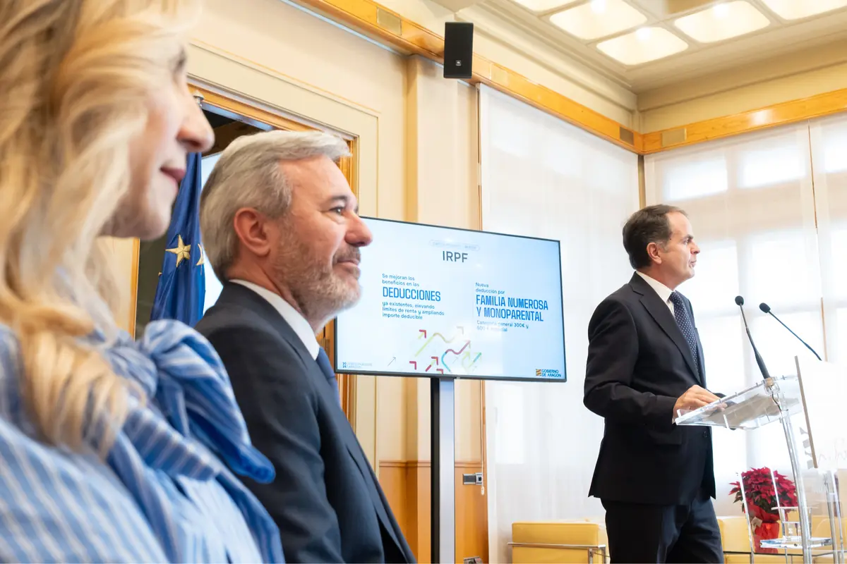 Tres personas en una sala de conferencias, una de ellas hablando en un atril mientras las otras dos escuchan, con una pantalla al fondo mostrando información sobre deducciones fiscales y familias numerosas
