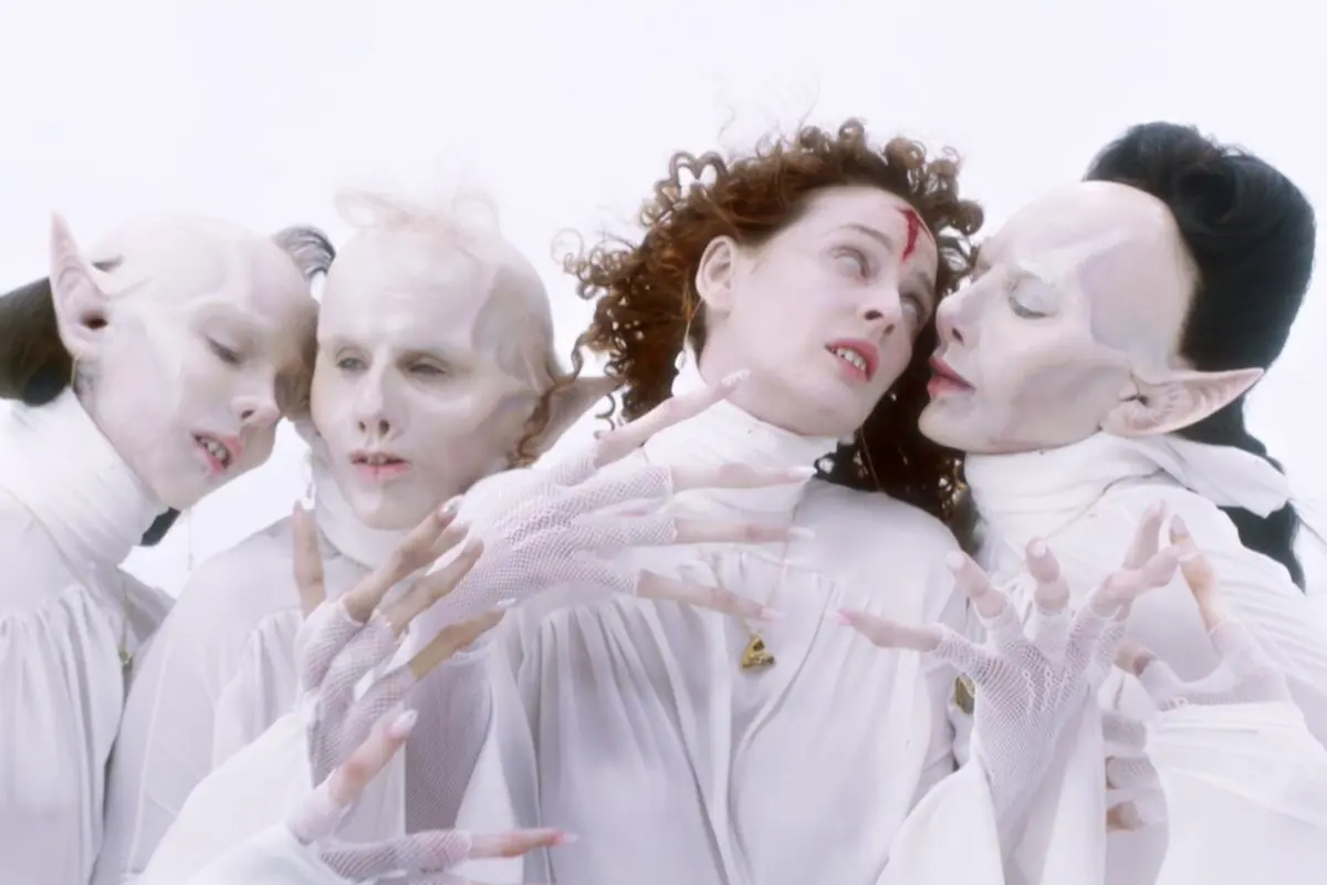 Cuatro personas con maquillaje y vestuario blanco de aspecto alienígena o fantástico posan juntas en un fondo claro, con expresiones dramáticas y manos extendidas. Cuatro personas con maquillaje y vestuario blanco de aspecto alienígena o fantástico posan juntas en un fondo claro, con expresiones dramáticas y manos extendidas.