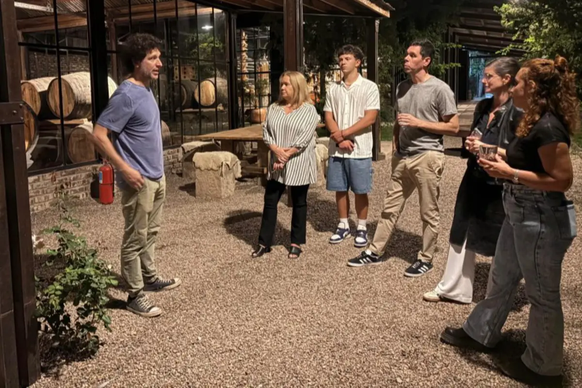 Un hombre habla frente a un grupo de cinco personas en un espacio al aire libre con barriles de madera al fondo