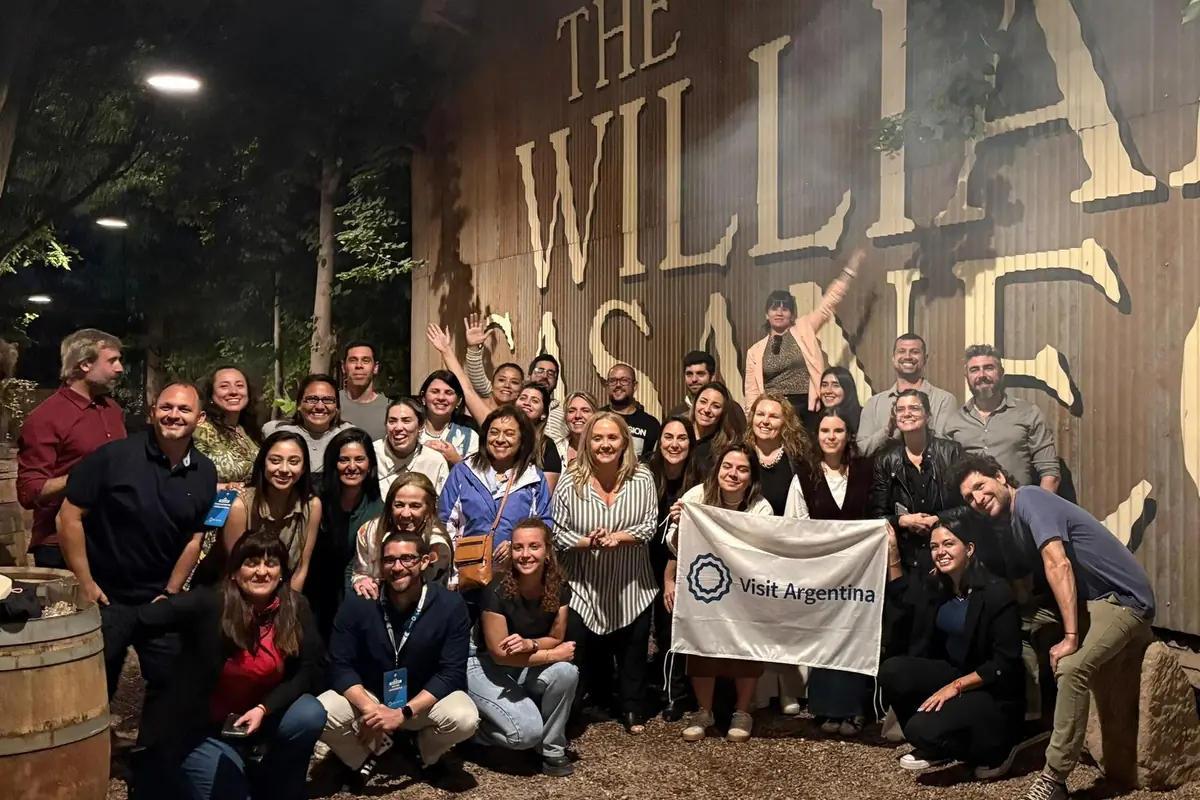 Grupo de personas posando sonrientes frente a un mural grande, algunas sostienen una bandera que dice Visit Argentina