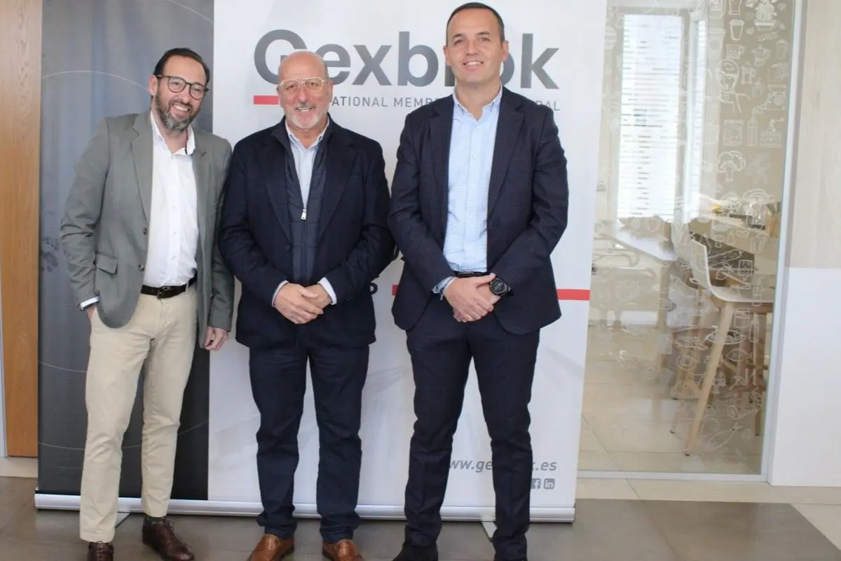 Tres hombres vestidos de traje posan sonrientes frente a un cartel de Gexbrok en una oficina moderna