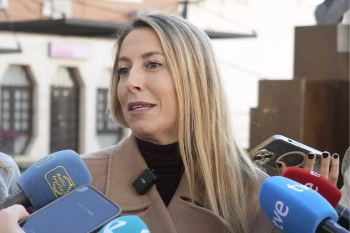 Mujer rubia con abrigo beige hablando al aire libre rodeada de varios micrófonos de prensa