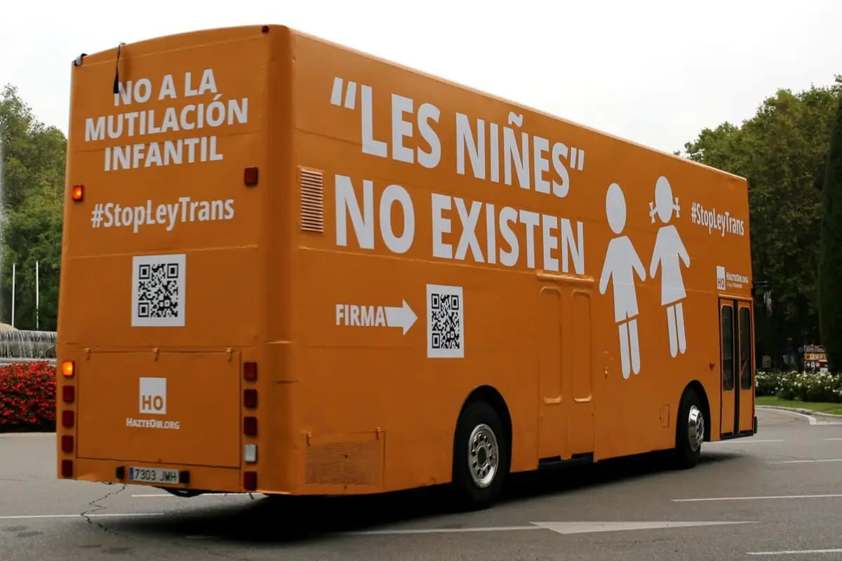 Autobús naranja con mensajes en contra de la ley trans y la identidad de género no binaria circulando por una calle
