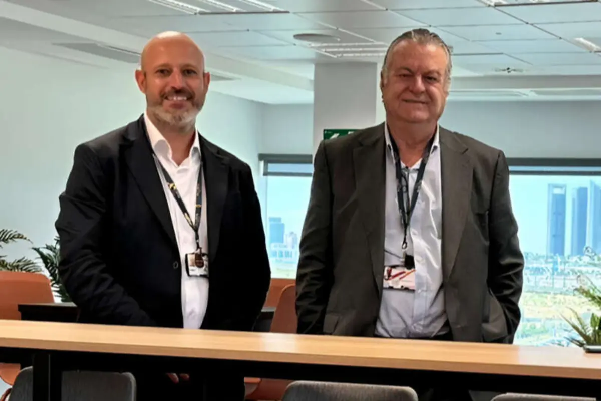 Dos hombres con traje y gafete posan sonrientes en una oficina moderna con ventanales y vista a la ciudad.