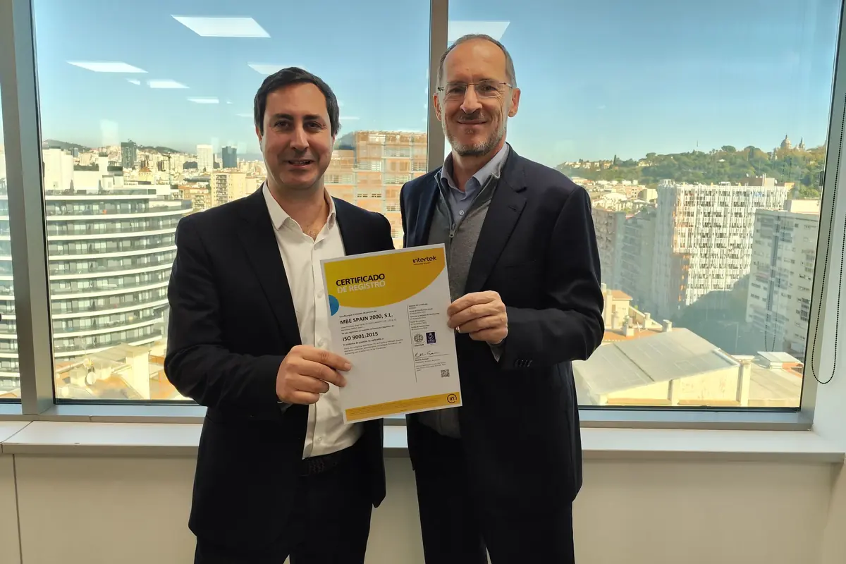 Dos hombres de traje sonríen mientras sostienen un certificado frente a una ventana con vista a una ciudad moderna y edificios altos