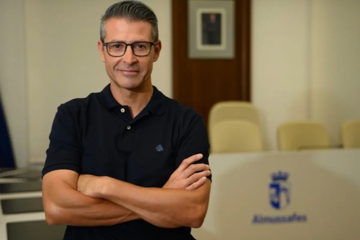 Hombre de cabello corto y canoso con gafas y camiseta polo negra posando con los brazos cruzados en una sala de reuniones con sillas beige y un escudo azul al fondo