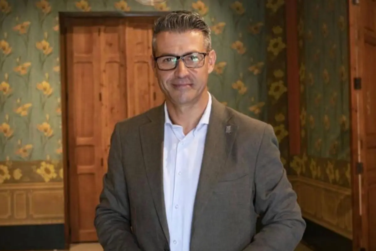 Hombre de cabello canoso y gafas con saco marrón y camisa blanca posando en una habitación con paredes decoradas con flores amarillas Hombre de cabello canoso y gafas con saco marrón y camisa blanca posando en una habitación con paredes decoradas con flores amarillas