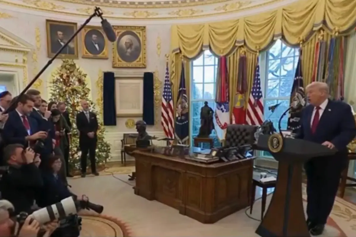 Un grupo de periodistas y fotógrafos cubre una conferencia en la Oficina Oval decorada con un árbol de Navidad mientras una persona de traje habla desde un podio junto al escritorio presidencial.