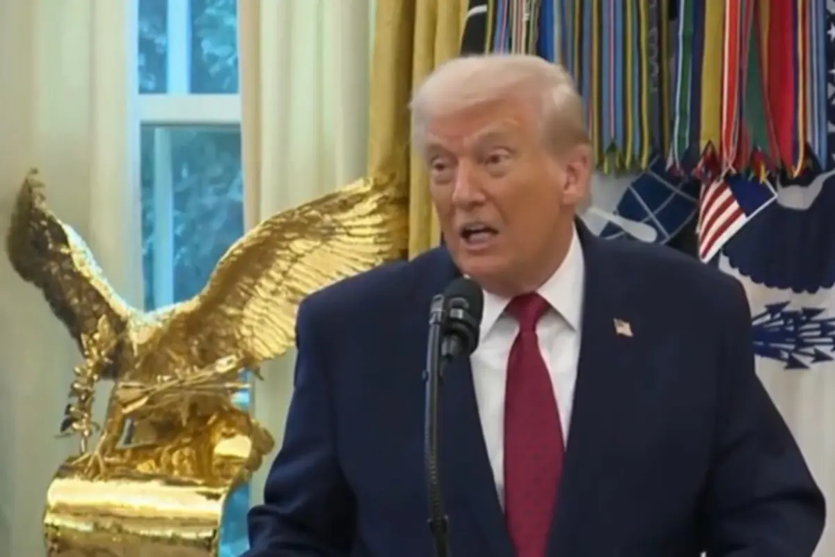 Hombre de cabello rubio con traje azul y corbata roja hablando frente a un micrófono en una sala decorada con una escultura dorada de águila y varias cintas de colores al fondo