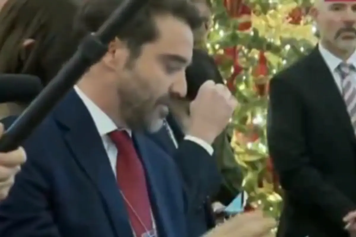 Hombre con barba y traje azul con corbata roja hablando frente a un micrófono con un árbol de Navidad decorado al fondo