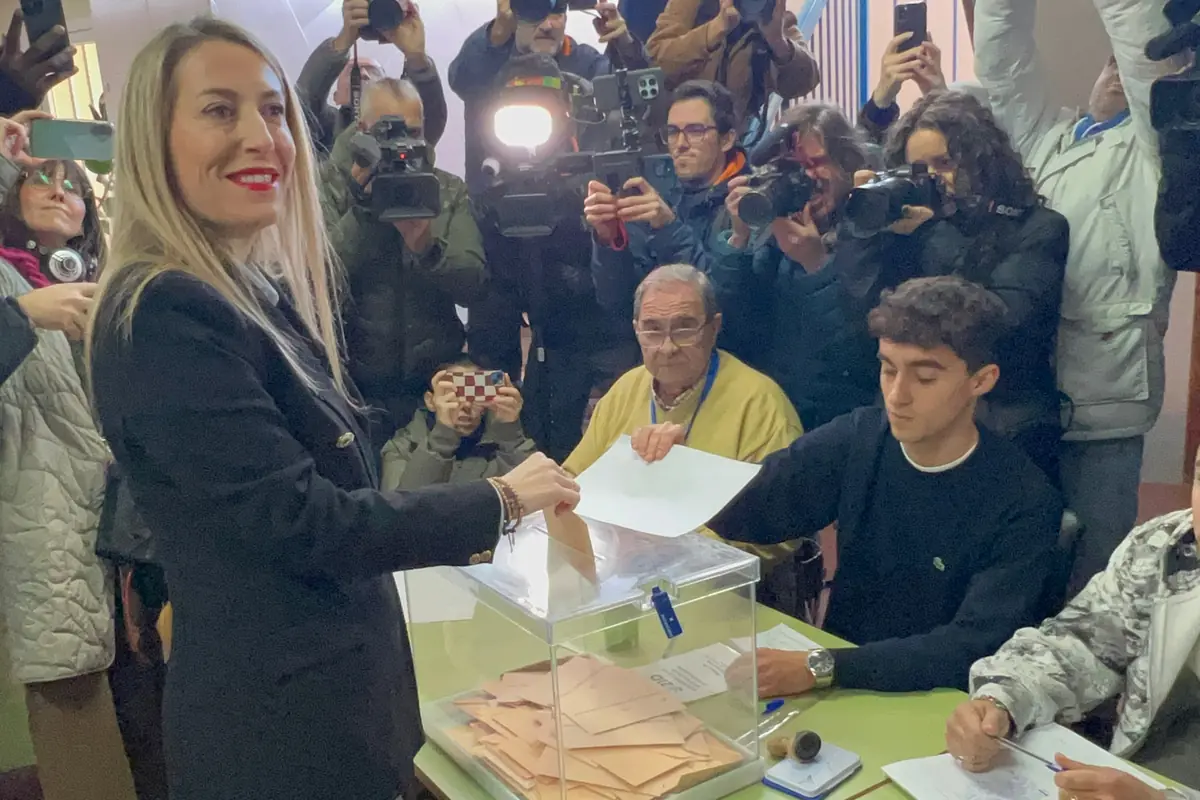 Mujer depositando su voto en una urna transparente en un colegio electoral rodeada de periodistas y fotógrafos que registran el momento