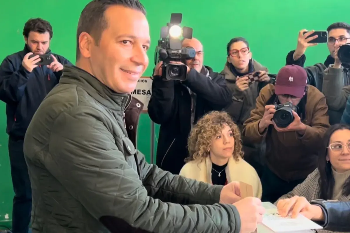 Hombre sonriendo deposita una papeleta en una mesa mientras varios periodistas y fotógrafos lo rodean y lo graban en un recinto con pared verde Hombre sonriendo deposita una papeleta en una mesa mientras varios periodistas y fotógrafos lo rodean y lo graban en un recinto con pared verde