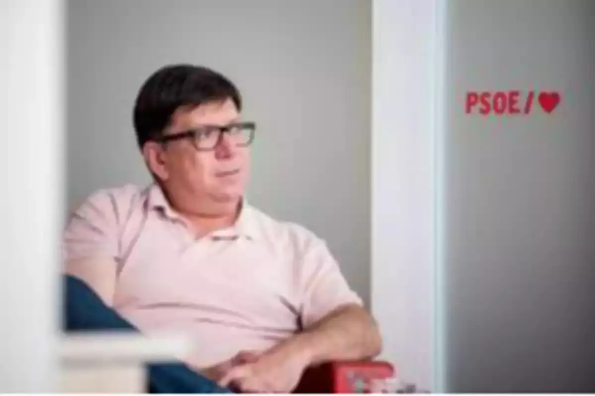 Hombre con gafas y camiseta clara sentado en una silla junto a una pared blanca con el texto PSOE y un corazón rojo Hombre con gafas y camiseta clara sentado en una silla junto a una pared blanca con el texto PSOE y un corazón rojo