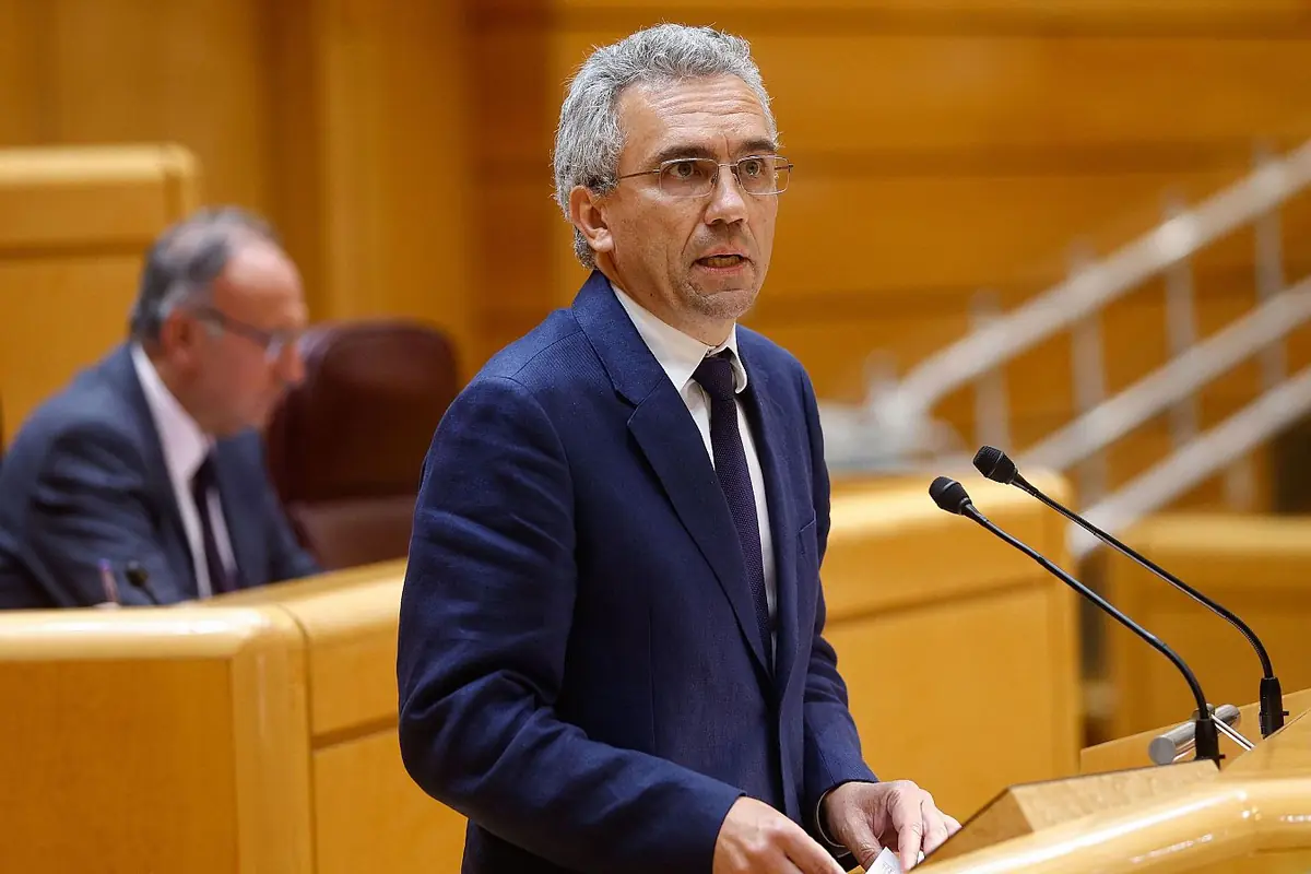 Hombre de cabello canoso y gafas hablando en un podio en un recinto parlamentario