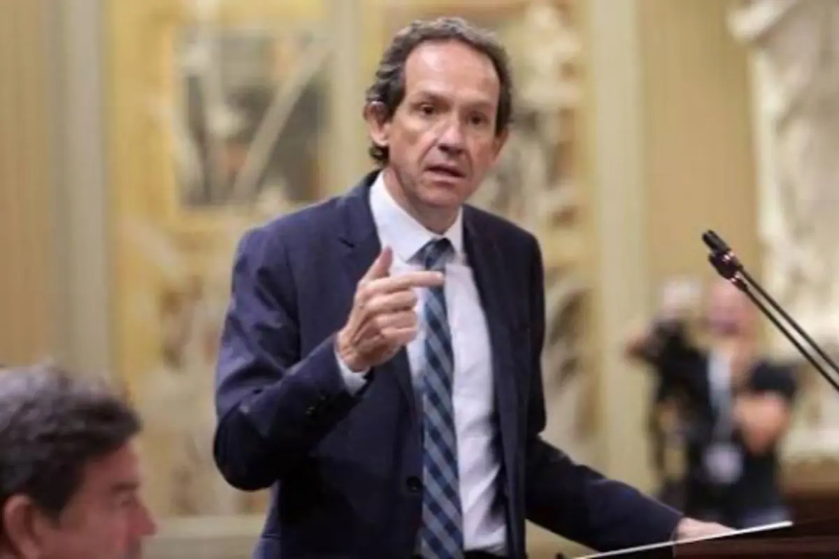 Hombre de mediana edad con traje y corbata hablando y gesticulando en un podio dentro de un salón formal