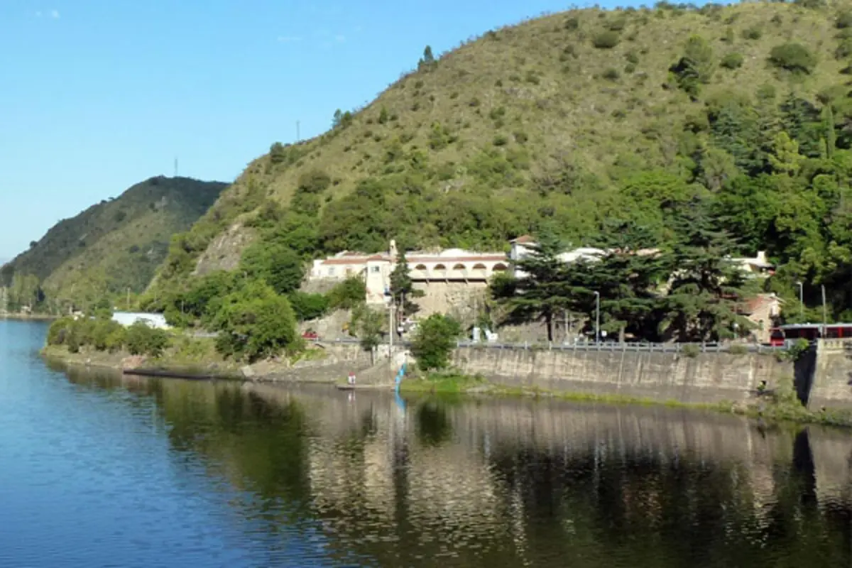 Vista de un lago tranquilo junto a una ladera verde con casas y un muro de contención en la orilla Vista de un lago tranquilo junto a una ladera verde con casas y un muro de contención en la orilla
