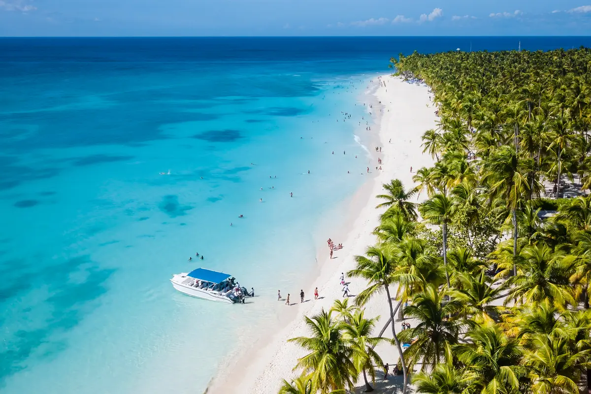 Playa de arena blanca con aguas turquesas, palmeras y una lancha cerca de la orilla donde varias personas disfrutan del mar