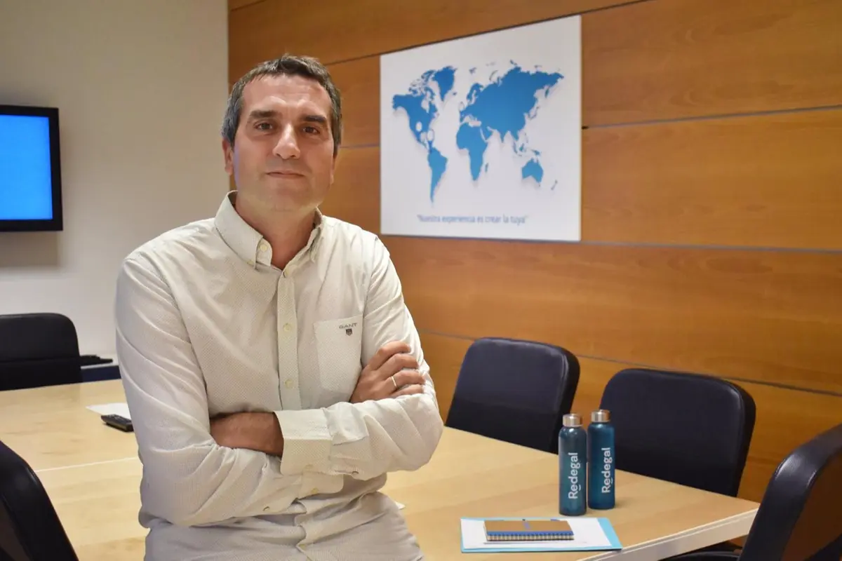 Hombre de cabello corto y camisa clara sentado en una sala de reuniones con una mesa, sillas, dos botellas azules y un cuaderno, al fondo un mapa del mundo en la pared