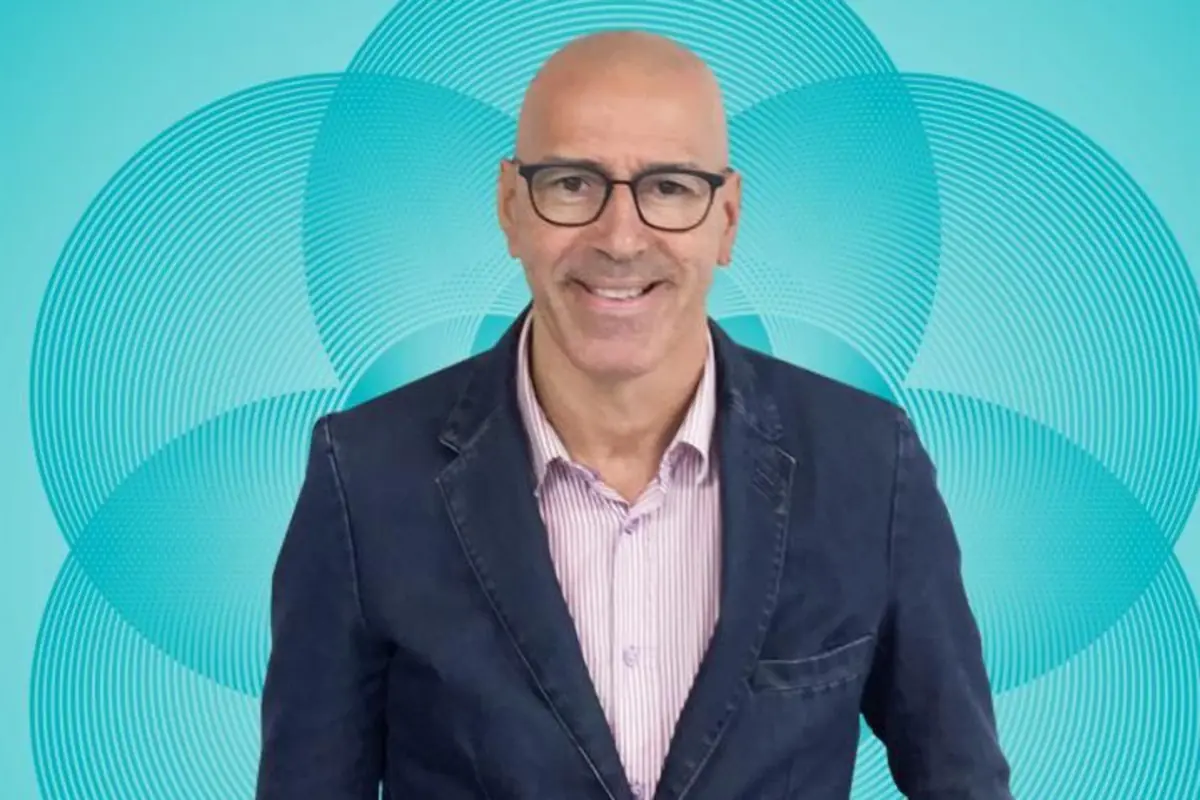 Hombre calvo con gafas y chaqueta azul sonriendo frente a un fondo turquesa con círculos concéntricos