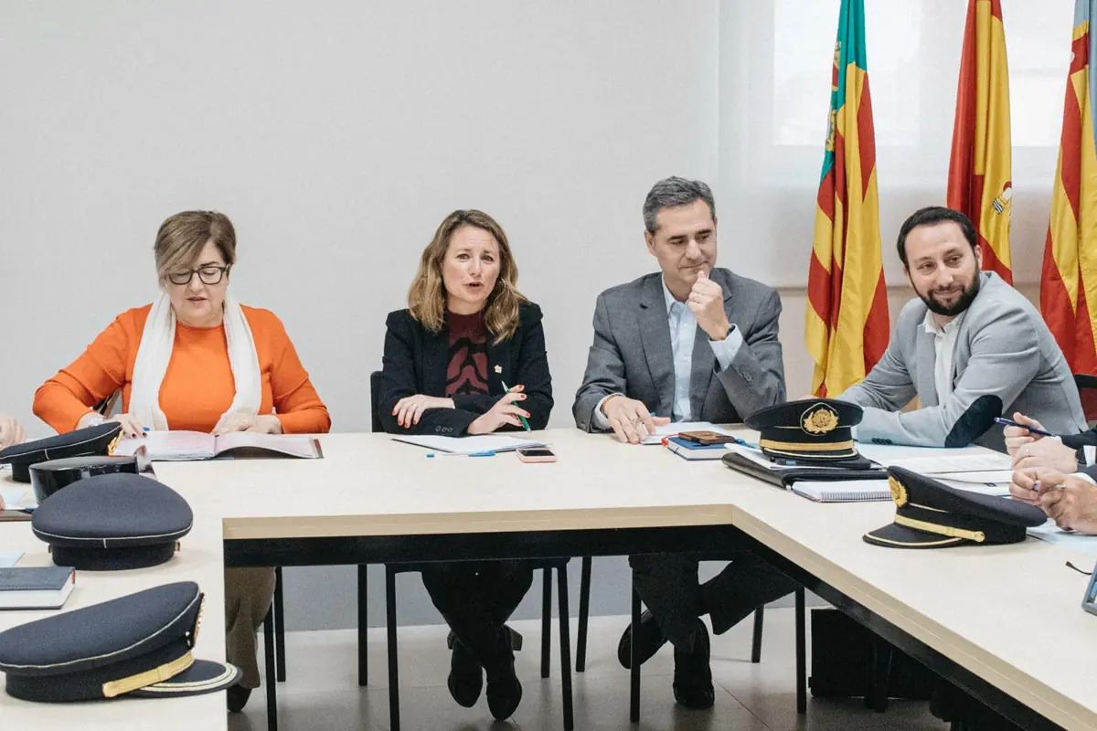 Reunión formal con cuatro personas sentadas en una mesa rodeadas de carpetas y gorras de uniforme con banderas oficiales al fondo