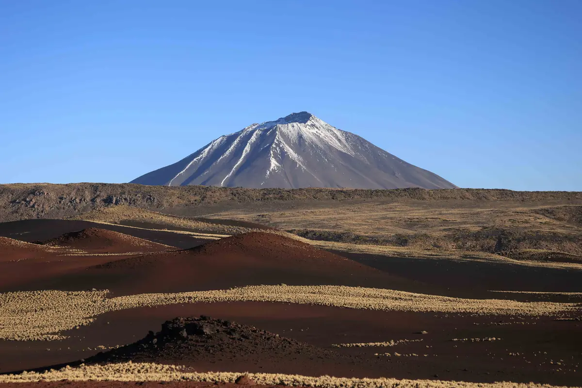 Un volcán nevado se eleva sobre un paisaje árido con colinas de tierra oscura y vegetación dispersa bajo un cielo despejado.