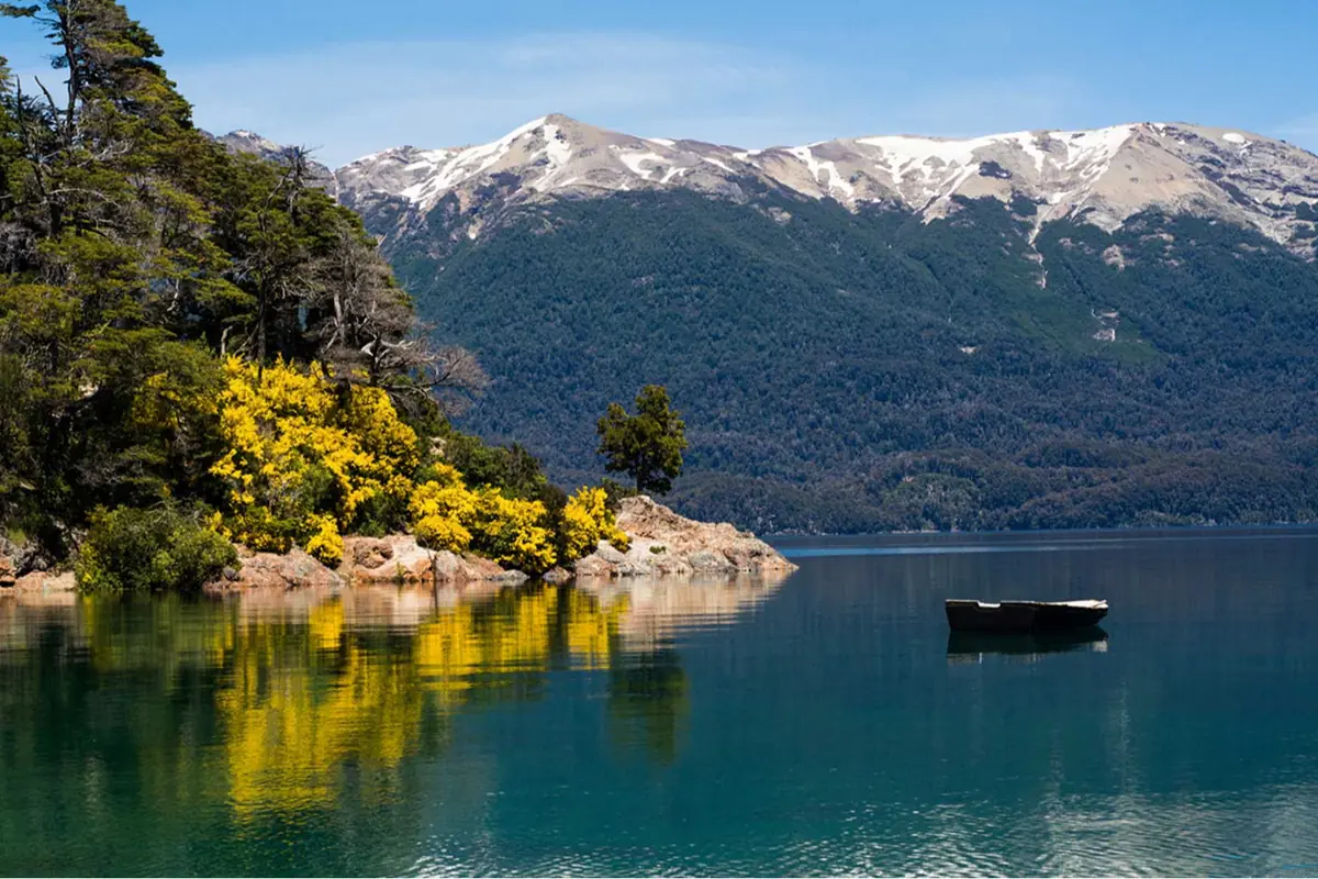 Un lago de aguas tranquilas con un bote, rodeado de montañas nevadas y árboles con flores amarillas reflejadas en el agua
