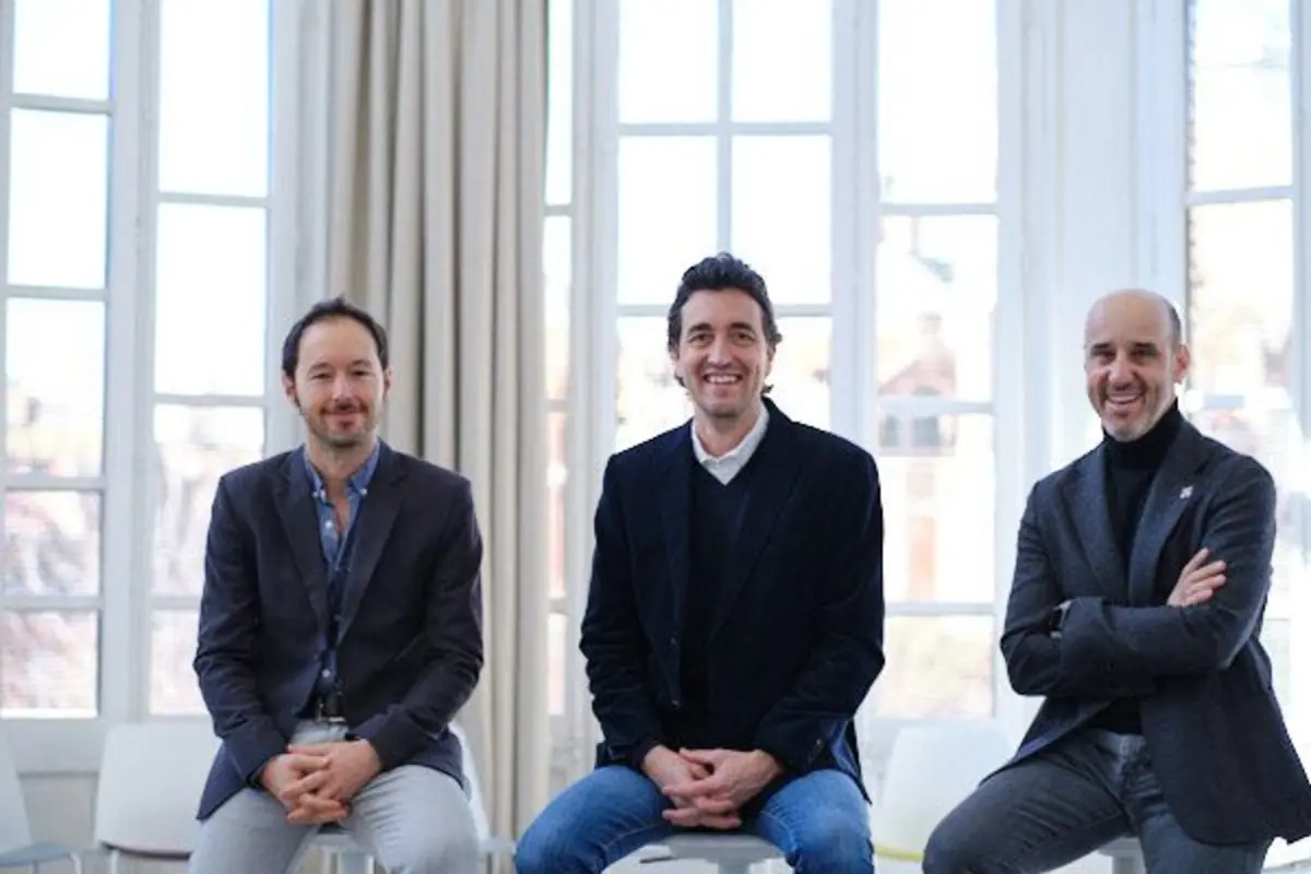 Tres hombres sonrientes con ropa formal casual sentados en sillas frente a grandes ventanales en una sala luminosa