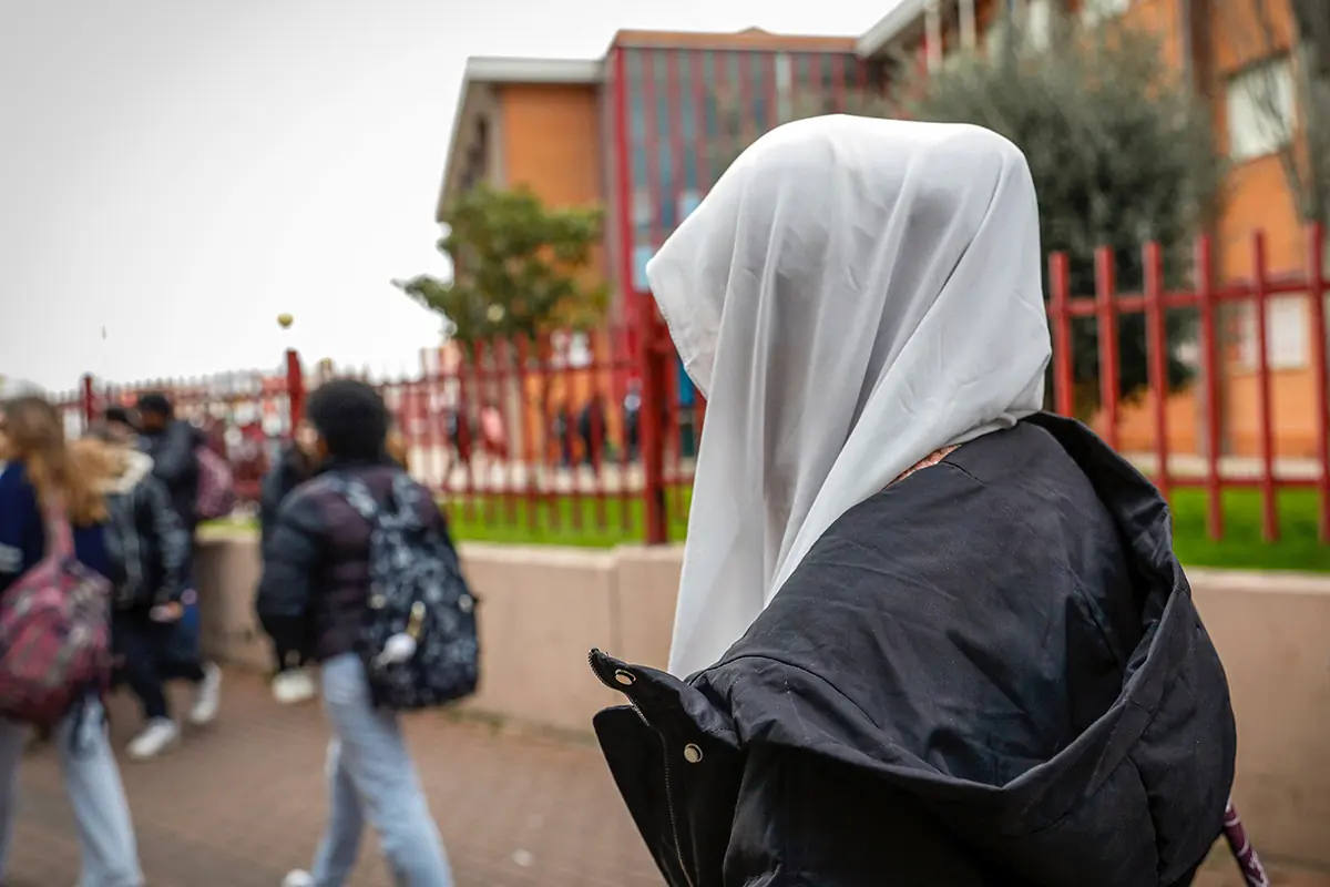 Una joven con velo blanco y chaqueta negra camina cerca de una escuela mientras otros estudiantes pasan a su alrededor
