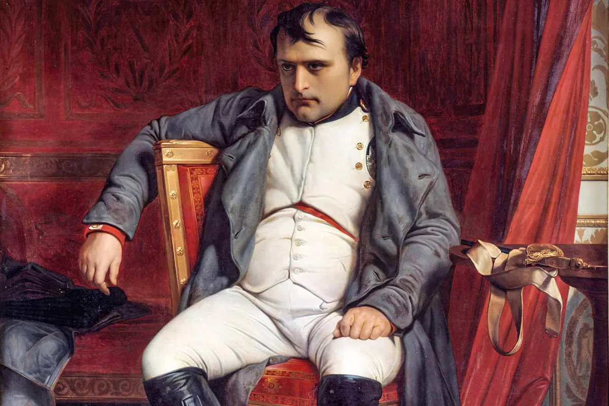 Hombre con uniforme militar blanco y capa gris sentado en una silla dorada con expresión seria en un entorno elegante de tonos rojos