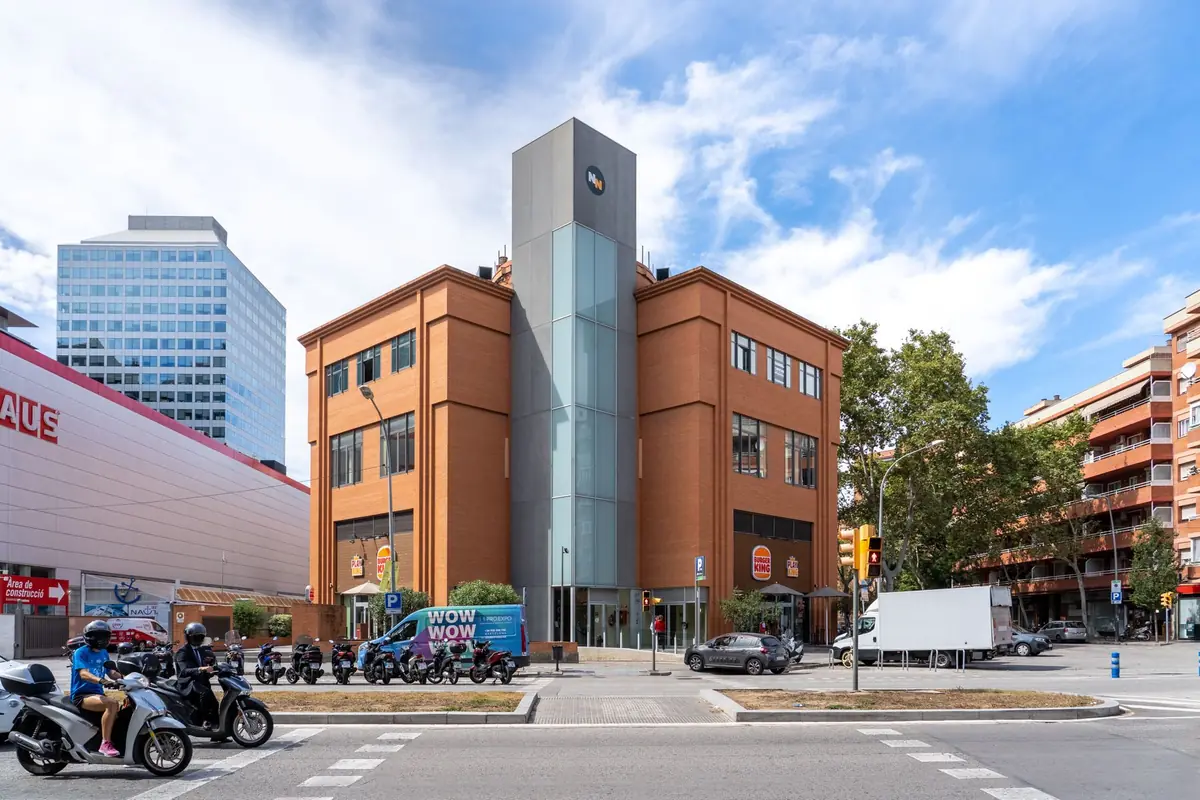 Edificio de ladrillo con una torre central de cristal en una esquina urbana, con un restaurante Burger King en la planta baja, motos y coches circulando y cielo parcialmente nublado.