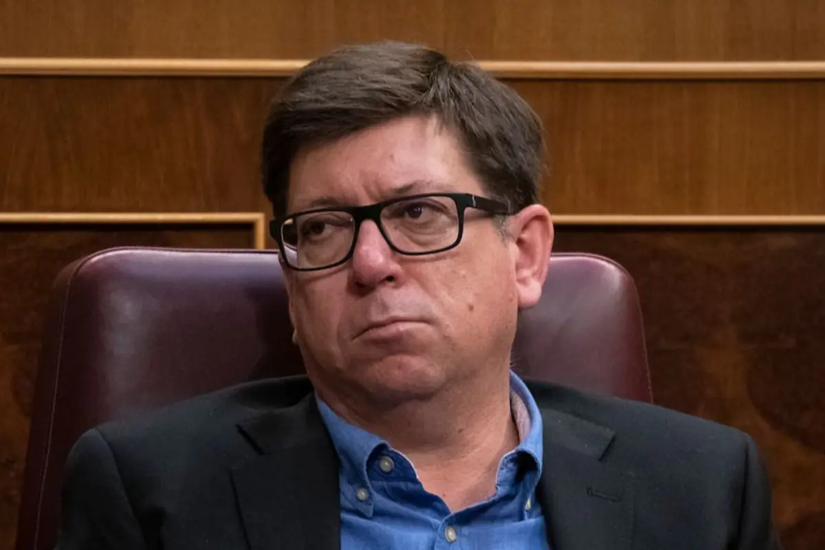 Hombre de cabello corto y gafas oscuras sentado en una silla de cuero marrón con expresión seria en un entorno de madera Hombre de cabello corto y gafas oscuras sentado en una silla de cuero marrón con expresión seria en un entorno de madera