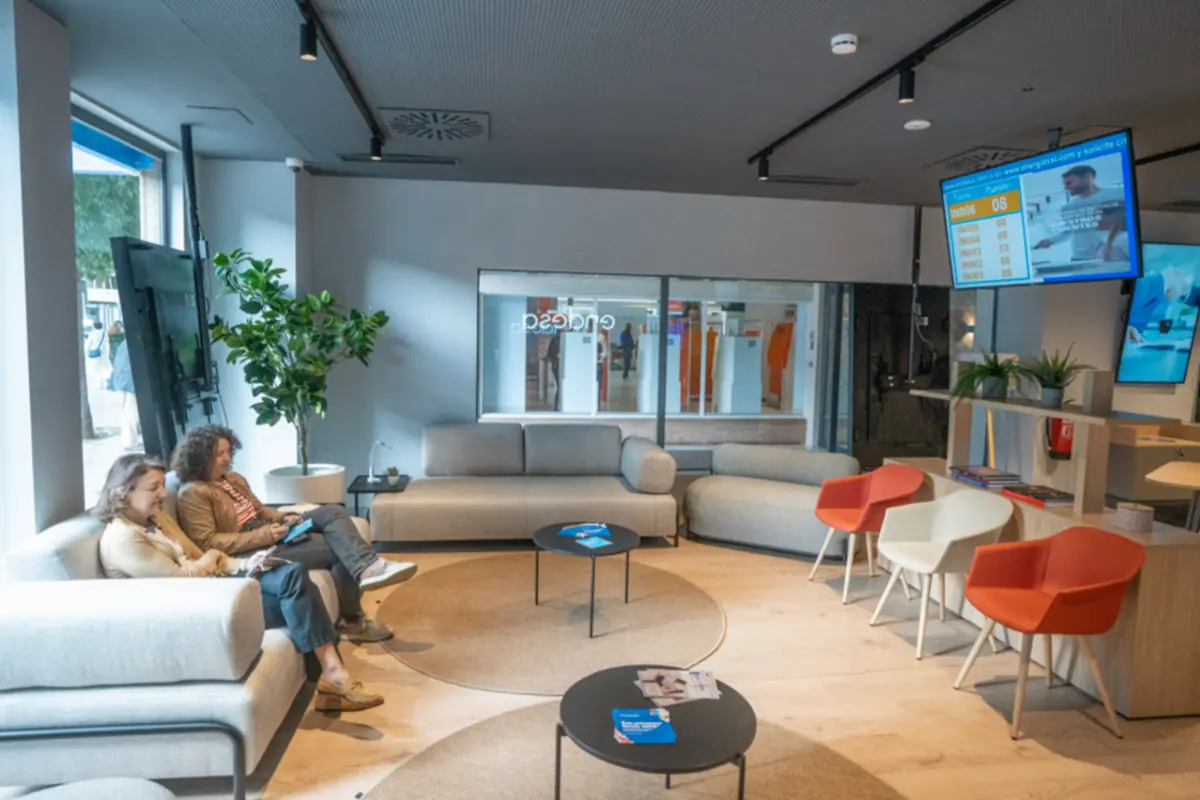 Dos mujeres sentadas en un sofá dentro de una sala de espera moderna con sillones grises, sillas de colores, mesas bajas, plantas y pantallas en la pared