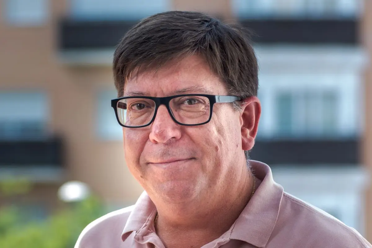 Retrato de un hombre de mediana edad con gafas negras y polo rosa sonriendo levemente al aire libre con edificios desenfocados de fondo