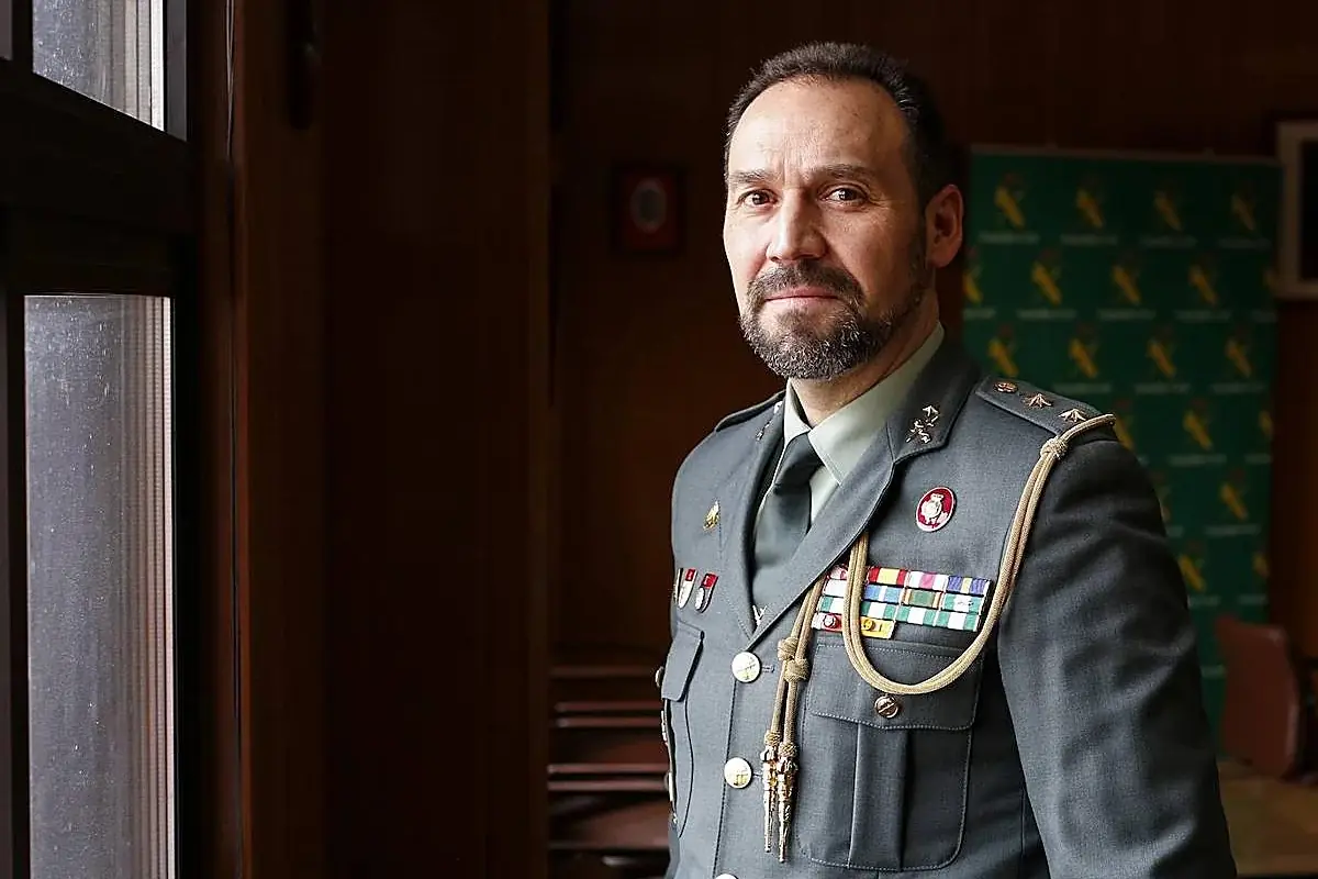 Hombre con barba y uniforme militar con varias medallas y condecoraciones de pie junto a una ventana en una habitación iluminada suavemente