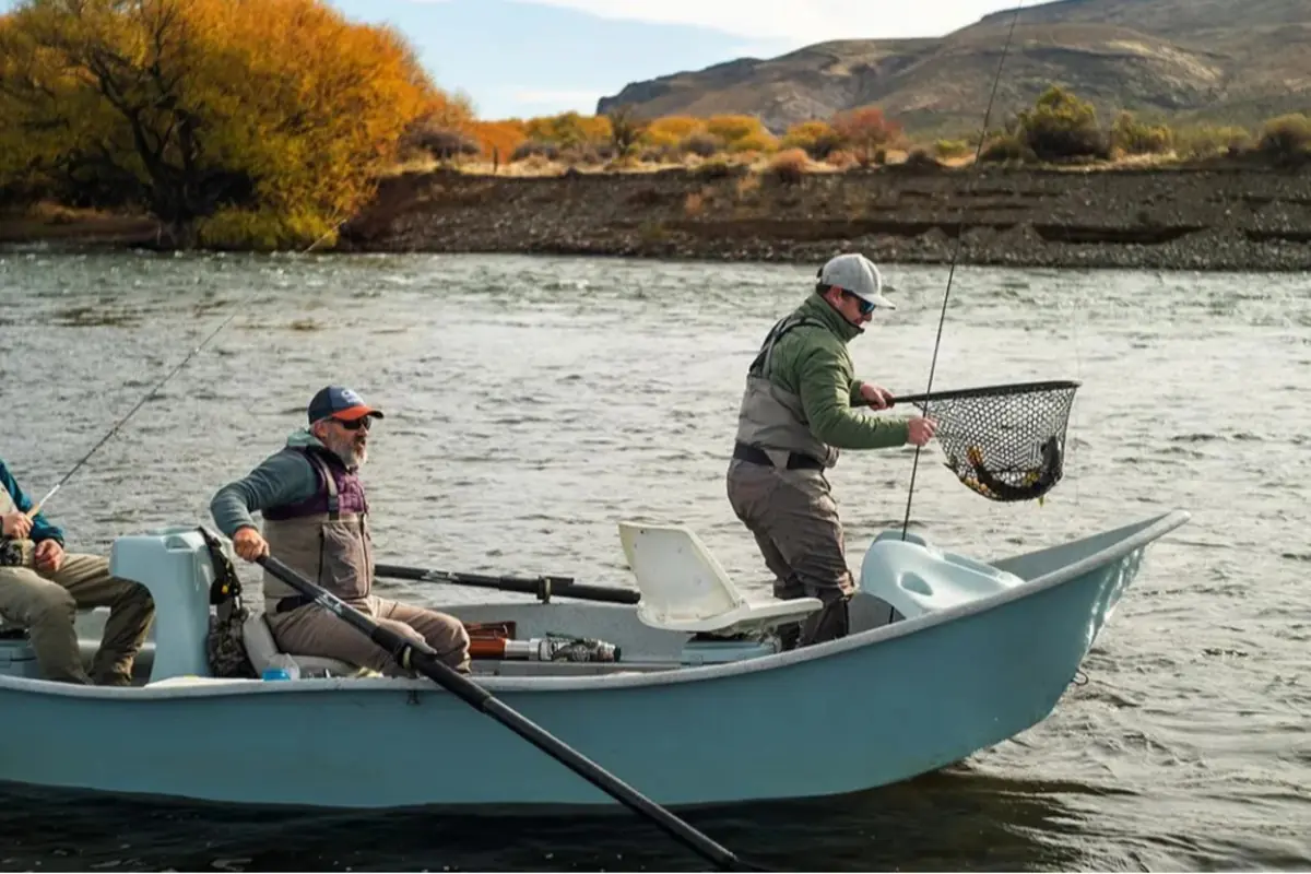 Tres personas pescando en un bote sobre un río, una de ellas sostiene una red con un pez mientras otra rema y al fondo se ven árboles con hojas otoñales y montañas.