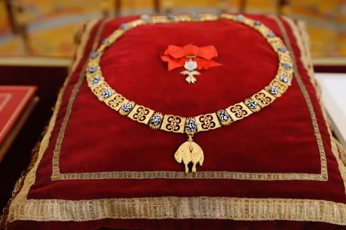 Collar dorado con detalles azules y una figura de carnero al centro colocado sobre un cojín de terciopelo rojo con bordes dorados