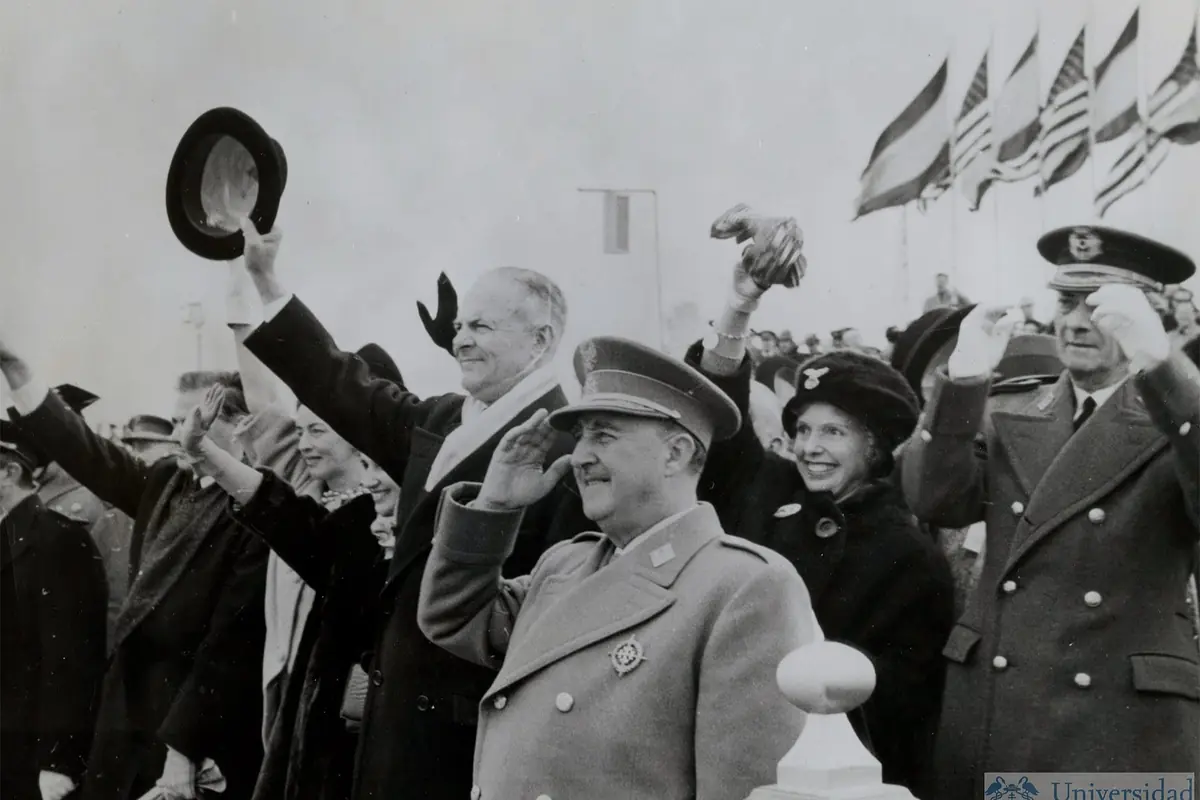 Francisco Franco en acto oficial al aire libre saludando y agitando sombreros mientras varias banderas ondean al fondo