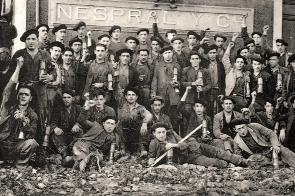 Fotografía histórica en blanco y negro de un gran grupo de mineros posando frente a un edificio con el letrero Nespral y Cía muchos de ellos levantan lámparas de mina y el puño en señal de unidad