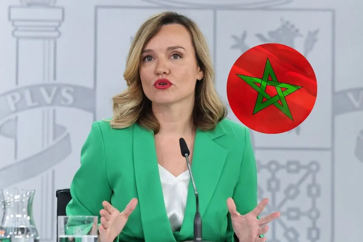 Mujer con saco verde hablando frente a un micrófono con un círculo que contiene la bandera de Marruecos a su lado