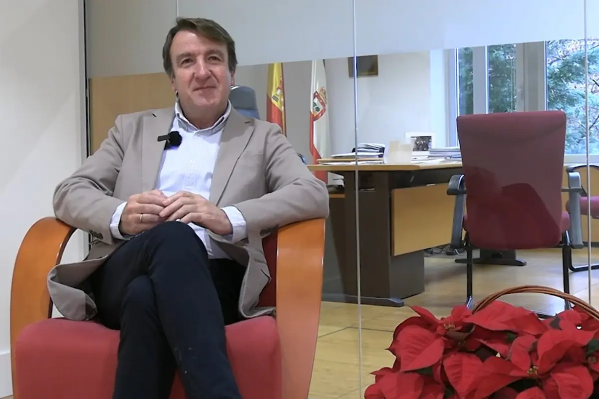 Hombre de mediana edad sentado en un sillón rojo en una oficina moderna con banderas al fondo y una planta de flores rojas en primer plano