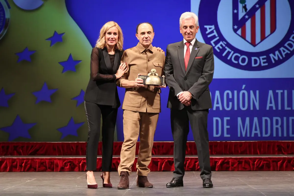 Tres personas posan en un escenario durante una ceremonia de premios del Atlético de Madrid mientras el hombre del centro sostiene un trofeo Tres personas posan en un escenario durante una ceremonia de premios del Atlético de Madrid mientras el hombre del centro sostiene un trofeo