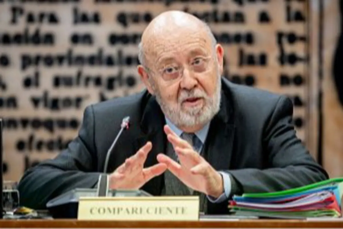 Hombre mayor con barba y gafas hablando en una mesa con un micrófono y documentos, detrás de un letrero que dice compareciente